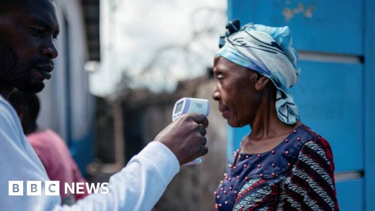 A man in a white shirt holding a contactless infrared thermometer near a woman's face, who is wearing a blue headscarf and patterned dress, amidst an Ebola outbreak in DR Congo.