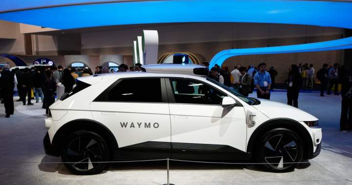 A Waymo Ojai driverless car displayed at a tech show, illuminated by bright lights.