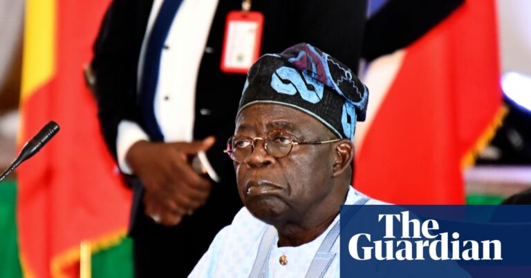 President Bola Ahmed Tinubu, in a traditional cap and glasses, sits at a desk with a microphone, wearing white robes, presiding over an event.