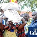 Ugandan woman carries grain bag, watched by dignitaries at a food distribution point in Karamoja.