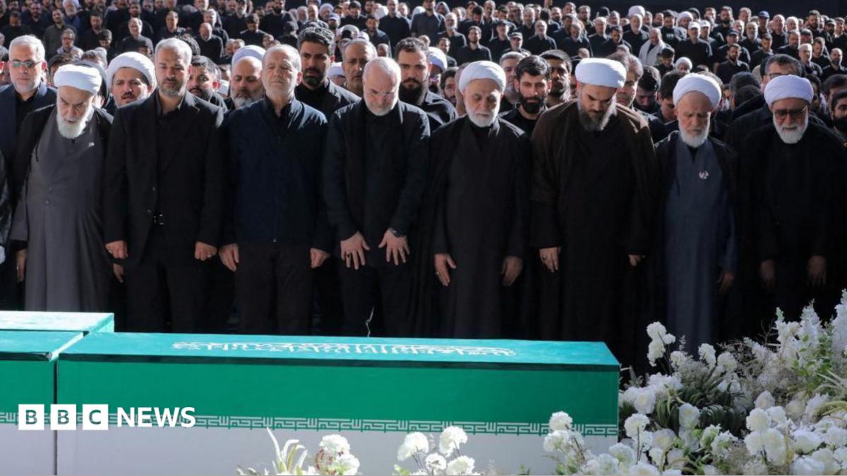 Image of thousands of mourners gathered at a funeral in Tehran, with senior Iranian officials in attendance, but Mojtaba Khamenei visibly absent.