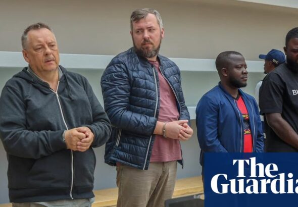 Four men in handcuffs standing behind a table with laptops in an Angolan courtroom during a hearing in August 2025. Two Russian men, Lev Lakshtanov and Igor Ratchin, were sentenced for terrorism and spying.