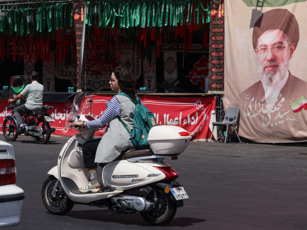 Image showing a woman riding a motorcycle past a banner of Iran's Supreme Leader Mojtaba Khamenei in Tehran, with the context of ongoing US Iran strikes.