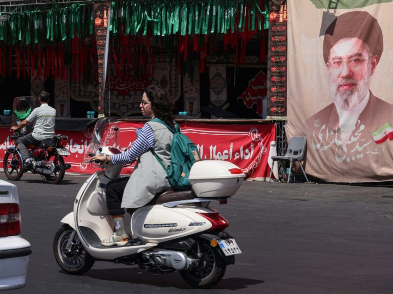 Image showing a woman riding a motorcycle past a banner of Iran's Supreme Leader Mojtaba Khamenei in Tehran, with the context of ongoing US Iran strikes.