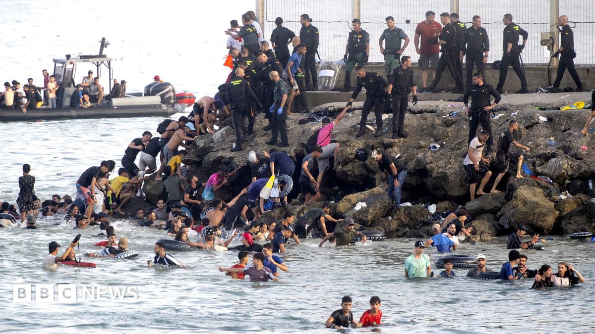 Aerial view of Ceuta with many migrants attempting to cross the border fence or swimming towards the shore.