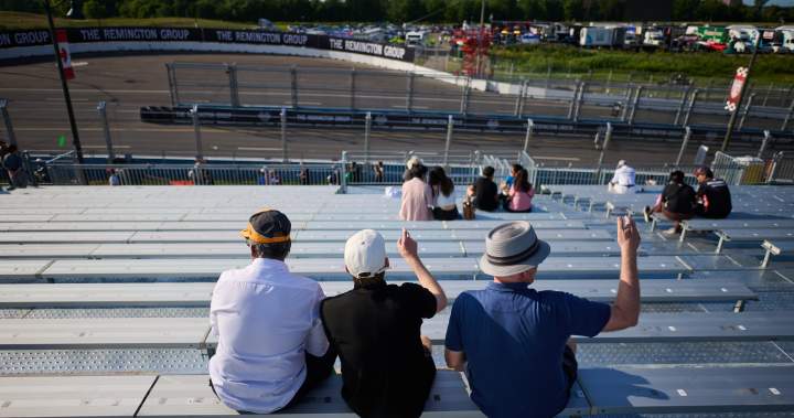 Aerial view of a vibrant red IndyCar vehicle speeding along a newly resurfaced street circuit in Markham, surrounded by temporary grandstands and excited crowds under a clear sky.