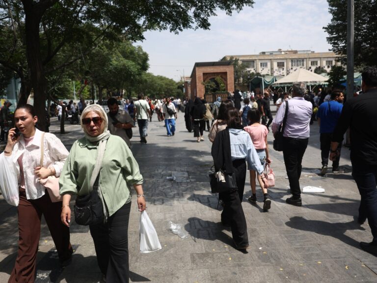 An Iranian man walks through the Grand Bazaar in Tehran, with blurry figures in the background. The atmosphere appears subdued.