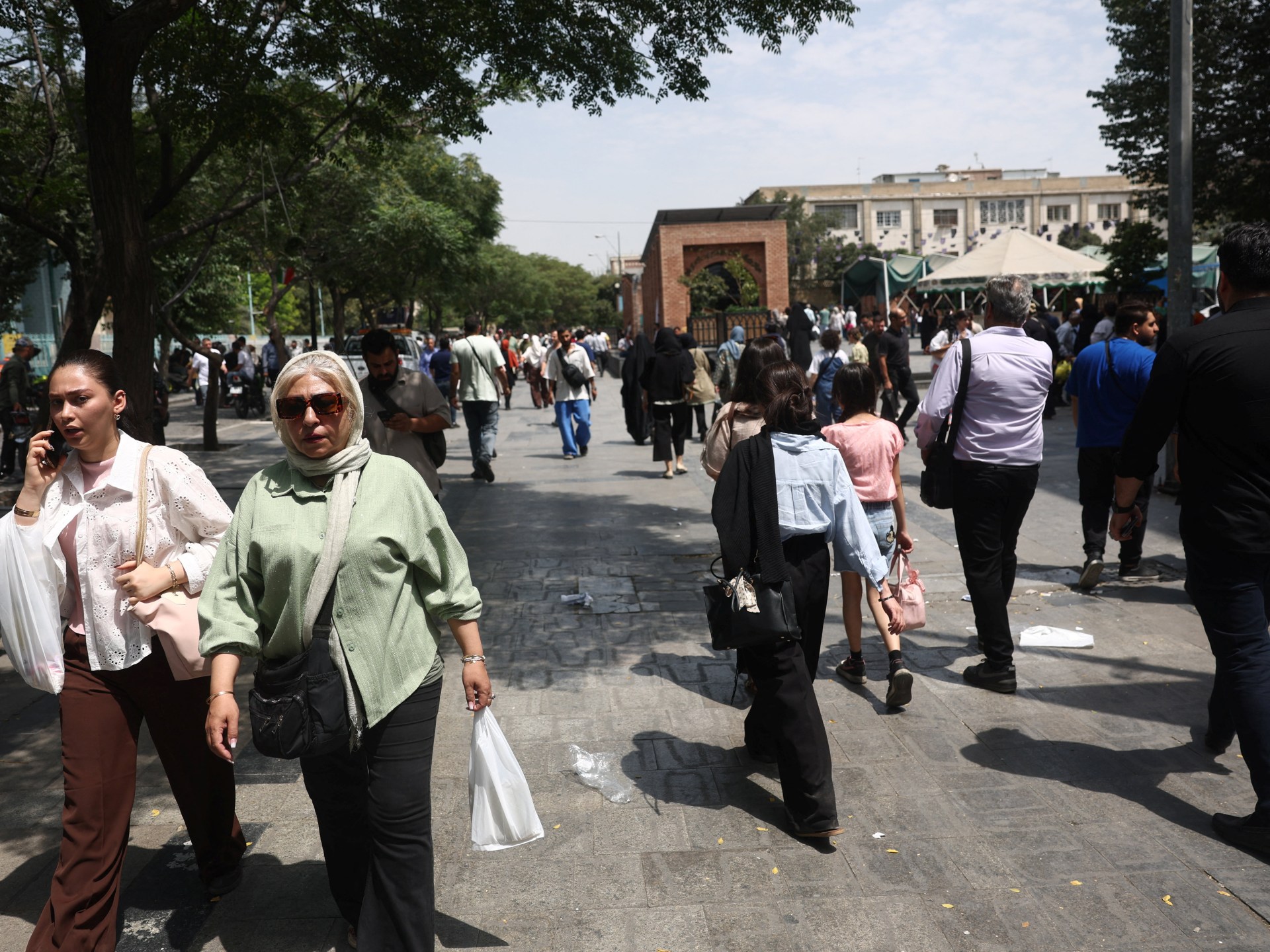 An Iranian man walks through the Grand Bazaar in Tehran, with blurry figures in the background. The atmosphere appears subdued.