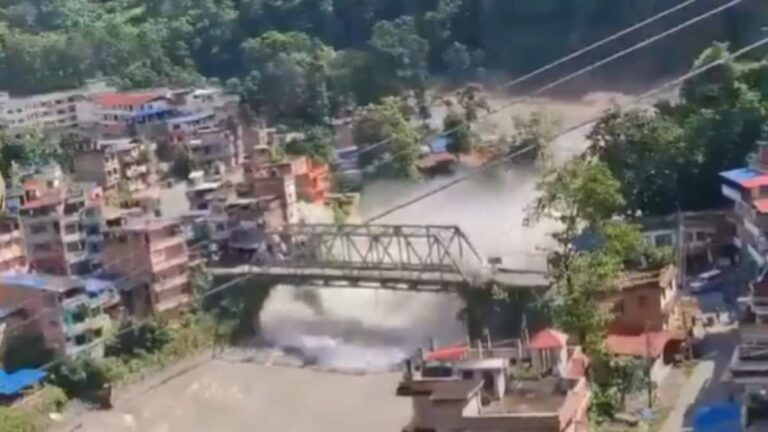 Aerial view of a swollen, muddy river in a mountainous region after a flash flood in Nepal, with rescue workers on the banks.