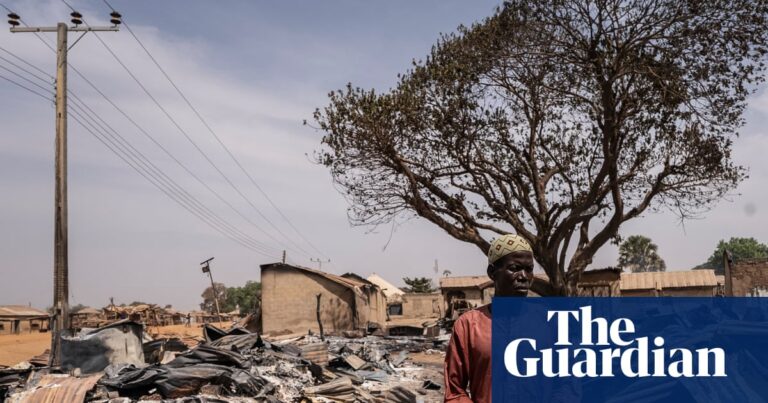 A man in a dark orange robe among burnt debris and damaged homes after an attack in Woro. Piles of ash and charred corrugated iron panels surround him.