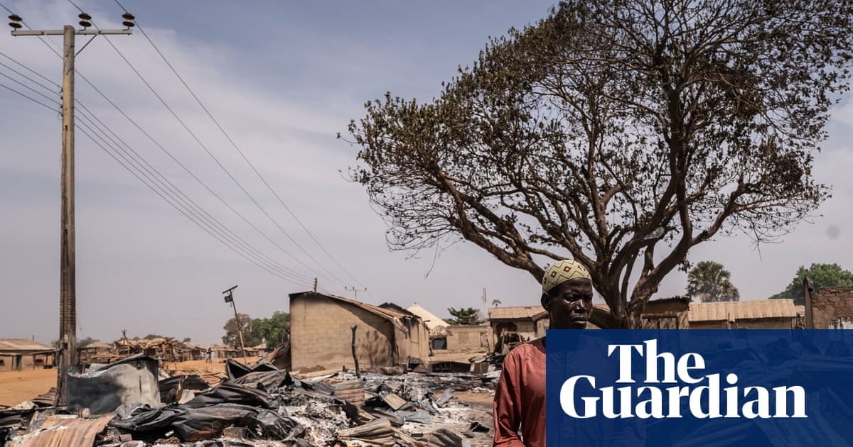 A man in a dark orange robe among burnt debris and damaged homes after an attack in Woro. Piles of ash and charred corrugated iron panels surround him.