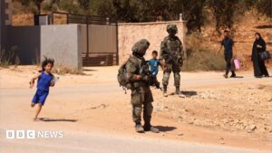 Image of Palestinian families walking on a dusty road with Israeli soldiers in the background, amidst reported displacements in Qusra, West Bank.