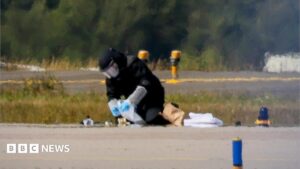 An image showing a blurred man defusing a bomb on an airport runway, symbolizing the ongoing Russian sabotage campaign in Europe.